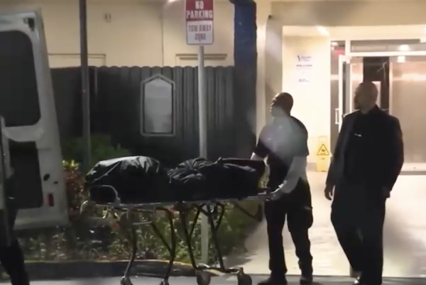 Officers in front of apartment building push cart with black body bag on top.