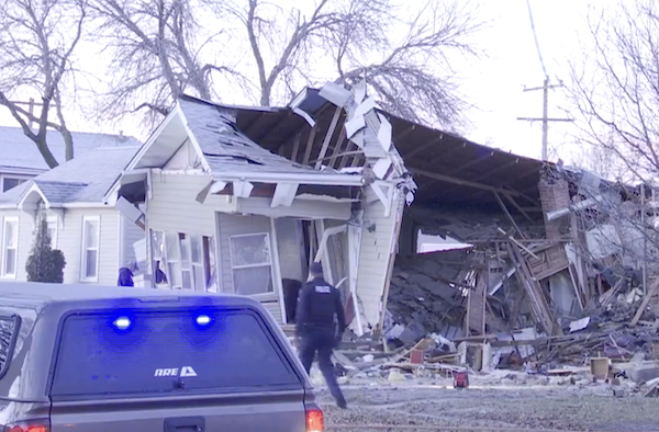 Light gray single story mid-century home on residential street is shown destroyed with the roof collapsed. A policeman in black walks past the front of the house and a police vehicle with lights flashing is on the street.