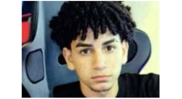 Teenager with black curly hair and thick eyebrows wearing a black shirt stares unsmiling at the camera.