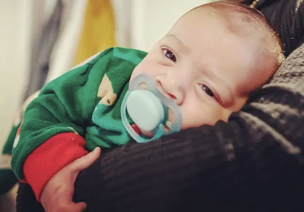 Photo of a baby with short brown hair and a pacifier in his mouth. He looks sleepy and wears a green and red shirt.