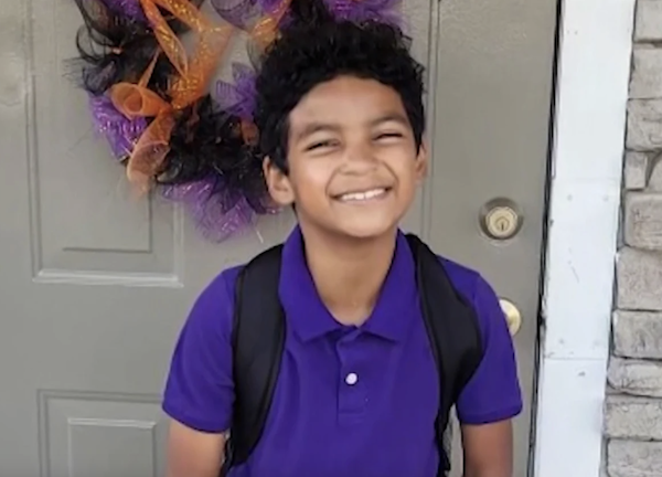 A little boy in a purple polo shirt wearing a backpack gives the camera a big smile. He stands outside the gray front door of a house that displays a colorful wreath. He has curly dark brown hair worn short.