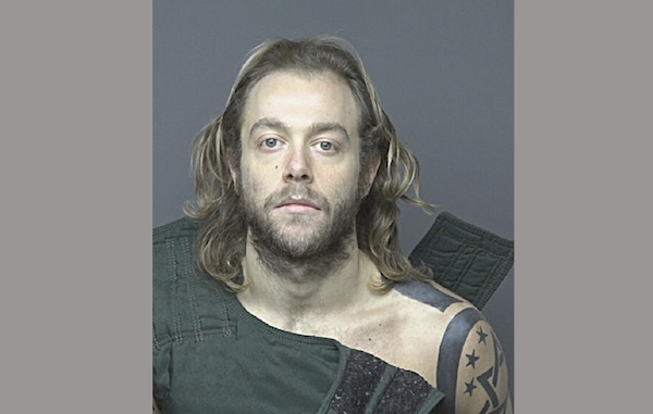 Mugshot of a man with straggling shoulder length medium blond hair and a full beard wearing a dark green shirt and showing a large shoulder tattoo with a star.