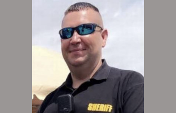 A white man with short medium brown hair wears dark sunglasses and a black shirt that says sheriff as he smiles at the camera.