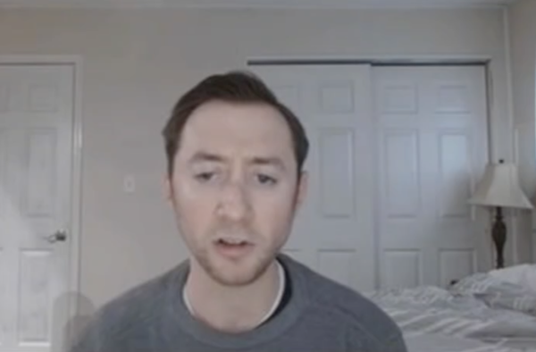 A white man with short brown hair and brown stubble beard sits on a bed in a suburban bedroom with white walls and closet doors. He wears a gray sweater and appears to be in the midst of speaking to the camera.