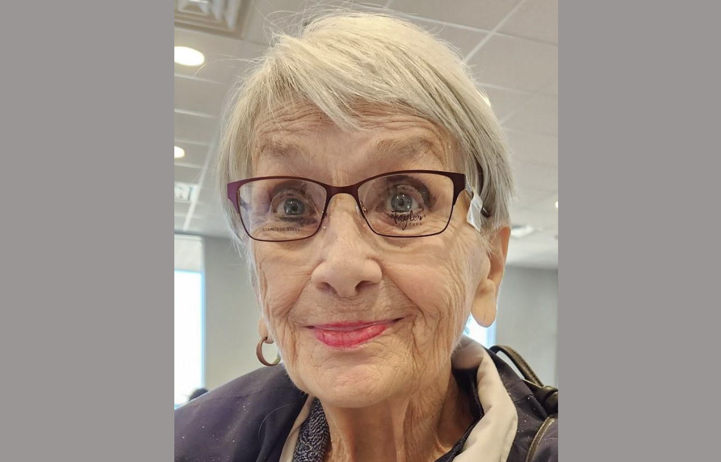 Daytime interior photo of a happy and energetic looking elderly lady with short gray hair and black-rimmed glasses.