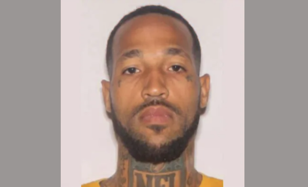 A man with short dark brown hair and a dark brown beard has neck and face tattoos. He frowns at the camera during his mugshot.