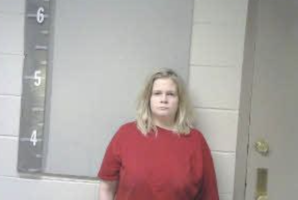 A white woman with disheveled long blonde hair wears a red t-shirt during her mugshot in a room with white cinder block walls.