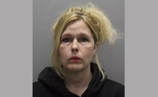 A middle-aged white woman with long blonde hair pulled up on her head with long bangs hanging down wears a black hoodie sweatshirt and bright red lipstick during her mugshot.