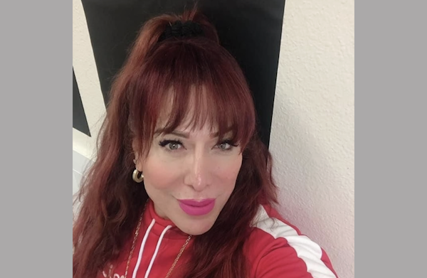 A middle-aged woman with long dark red hair wears a red track jacket and hoop earrings as she slightly smiles during a selfie.