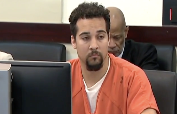 A young man with short curly dark brown hair and a dark brown goatee wears a jail-issued orange shirt as he sits at a table in a courtroom.