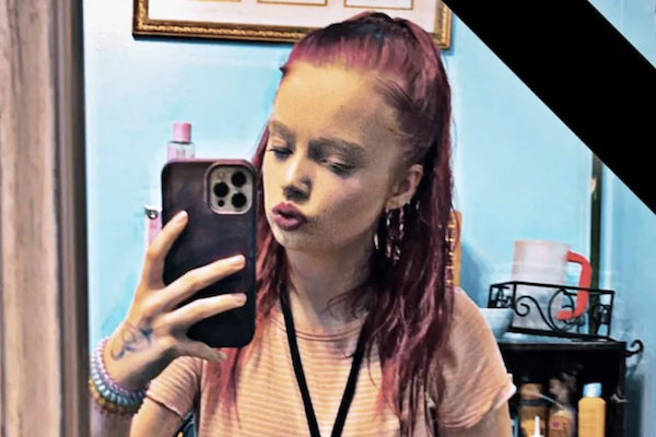A white teen girl with long pink-purple hair wears a light pink t-shirt and poses for a mirror selfie in a blue room.