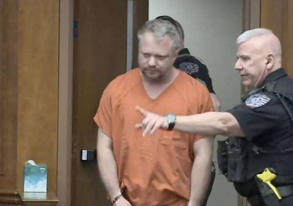 A middle-aged white man with a receding gray hairline and a brown beard is led into a courtroom by uniformed officers. He wears handcuffs and an orange jumpsuit.