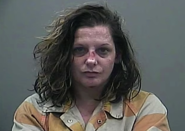 A disheveled middle-aged white woman with visible face wounds has wild shoulder length light brown hair and wears a white and orange striped shirt.