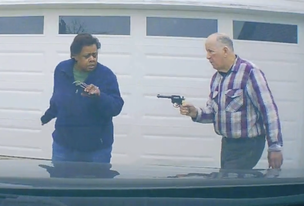 Daytime suburban driveway, footage from a vehicle dash camera facing the white garage door of a home. An older black woman with short dark brown hair on the left in a navy blue shirt turns away from an elderly white man with gray hair wearing a checked shirt who holds a revolver in his hand pointed at her.
