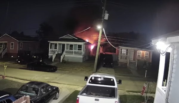 Security camera footage from a neighbor's house across a suburban street with older homes. A gray and white home has visible red flames rising from the rear.