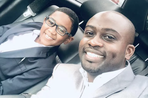 A man and boy in suits and dress shirts smile in the front seat of a car. The boy wears dark rimmed glasses.