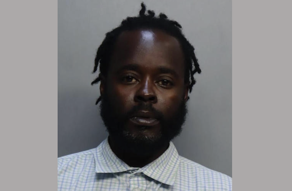 A man with unkempt dark brown locs and an full beard wears a light blue checked collared shirt during his mugshot.