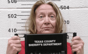 A white woman with shoulder-length straight gray-blonde hair frowns slightly during her mugshot against a height measurement on a white wall while she holds a black placard that reads: Texas County Sheriff's Department.
