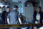 Nighttime scene looking through yellow crime scene tape. Two uniformed officers and a blonde civilian woman stand in the entrance of a suburban home's garage.