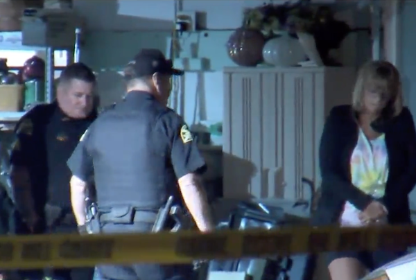 Nighttime scene looking through yellow crime scene tape. Two uniformed officers and a blonde civilian woman stand in the entrance of a suburban home's garage.
