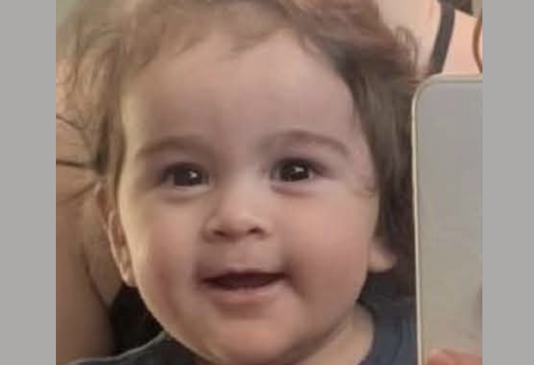A 1-year-old boy with light brown hair smiles while someone holds up a phone beside him to take a selfie.