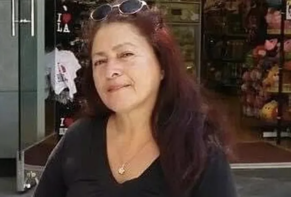 A woman with long dark red hair stands in front of a retail store on a sunny day. She wears a black shirt, a necklace, and sunglasses pushed up on her head as she tilts her head and slightly smiles at the camera.