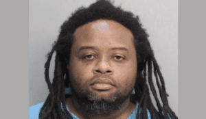 A man with small dreadlocks and a beard looks sleepy-eyed at the camera during his mugshot. He wears a blue polo shirt.
