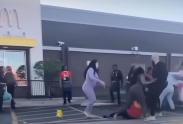 Daytime exterior parking lot view of a McDonald's. Several people including uniformed employees appear to be brawling beside the restaurant. A woman in a McDonald's uniform is on the ground as a man grabs her hair.