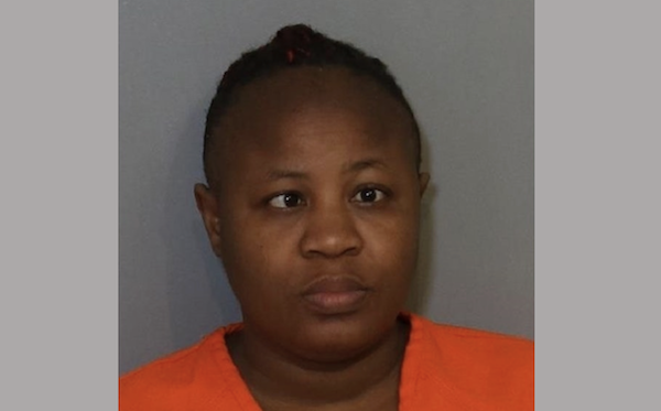 A black woman with dark brown hair pulled back in a high bun has crossed eyes. She stares expressionlessly forward and wears an orange shirt during her mugshot.