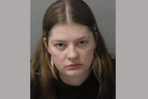 A young white woman with long, straight medium brown hair parted down the middle stares angrily at the camera during her mugshot. She wears a black shirt.