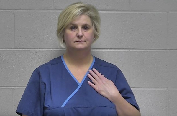 A middle-aged white woman in blue medical scrubs has bobbed light blonde hair and frowns slightly as she holds her left hand against her chest during her mugshot.