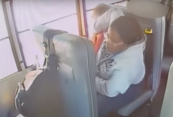 Overhead security camera footage of the interior of a school bus during daylight hours. A woman in a gray sweatshirt and glasses sits in a bus seat beside a young boy in an orange shirt and shoves him in the face.
