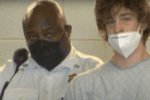 A young white man with unkempt overgrown short light brown hair wears a hospital gown and hospital mask pulled down below his nose. Beside him is a uniformed officer wearing a hospital mask.