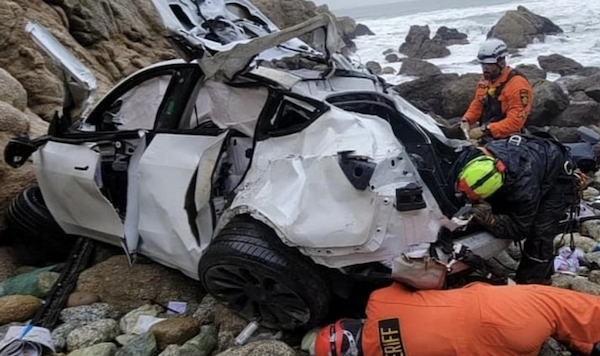 A mangled white Tesla sits on rocks at the foot of a cliff beside the ocean. Two emergency workers in orange and yellow examine the car.