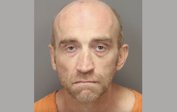 Mugshot of a white man with a bald pate and light brown sparse hair around his ears. He has light brown stubble on his face and looks depressed while he wears an orange shirt.