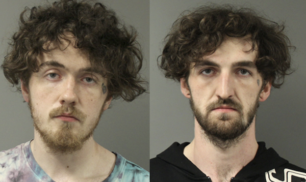 Side-by-side mugshots of two white men with brown curly hair worn at medium length around their ears. On the left, the man has a sparse light brown beard and wears a light blue tie-dyed t-shirt as he frowns. On the right, the man wears a black shirt and has a full brown beard while his expression is slack.