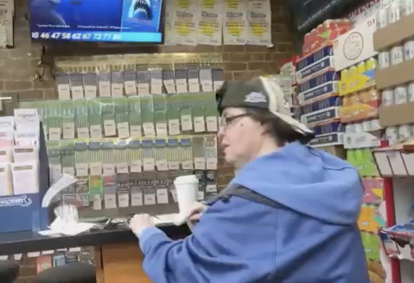 A woman in a black and white baseball cap and blue jacket has short brown hair and wears black rimed glasses. She sits at a counter ina convenience store with a cup of coffee.
