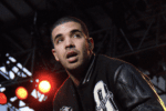 Drake pauses with mouth open while onstage during a concert. He has very short hair and wears a black letterman's jacket.