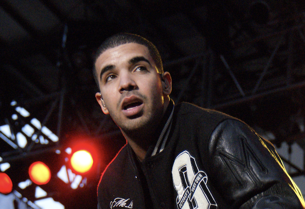 Drake pauses with mouth open while onstage during a concert. He has very short hair and wears a black letterman's jacket.