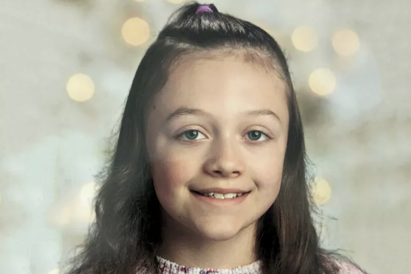 A pre-teen white child with long medium-brown hair pulled half up in a ponytail atop her head smiles with sad eyes during a formal portrait.