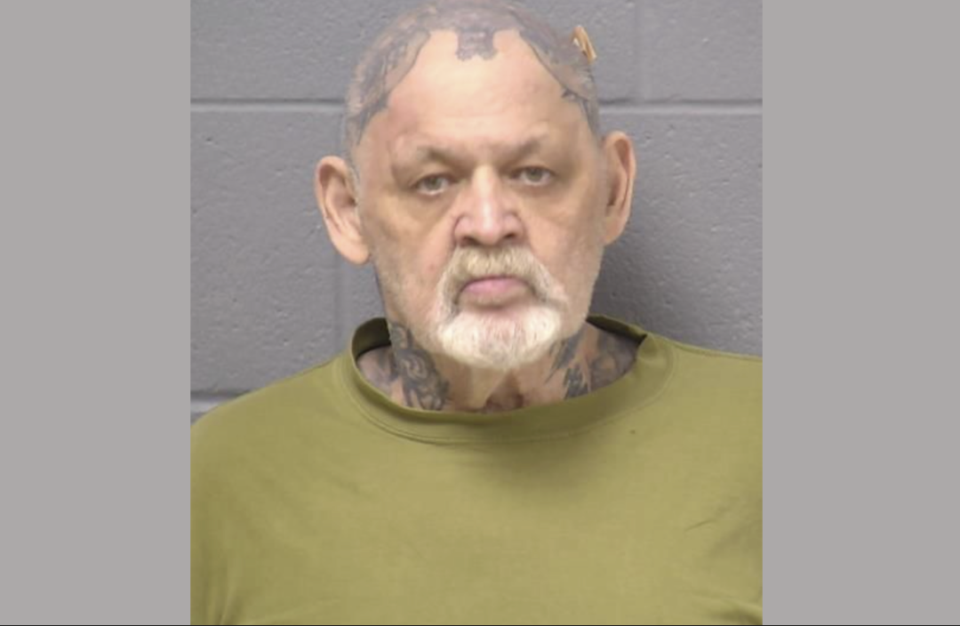 An elderly, bald white man with a gray goatee and mustache has many tattoos littering his neck and skull. He wears a green t-shirt and glares at the camera during his mugshot.