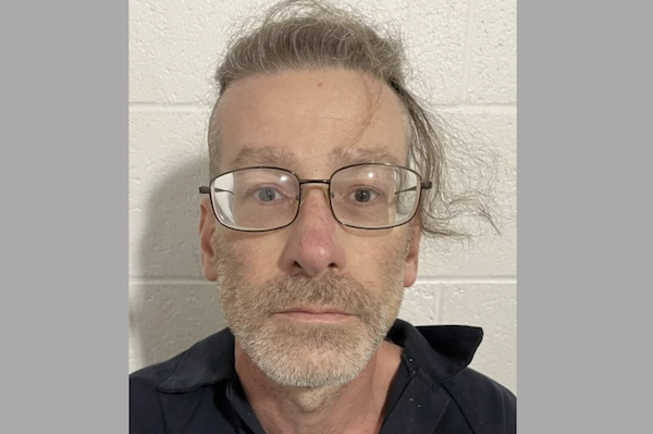 A middle-aged white man with graying disheveled hair wears black wire-rimmed glasses and has a full gray beard. He looks surprised during his mugshot as he wears an off-kilter dark collared shirt.