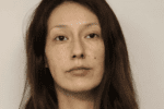 A young woman with long brown hair and a mole about one inch above the left corner of her mouth stares expressionless during her mugshot.