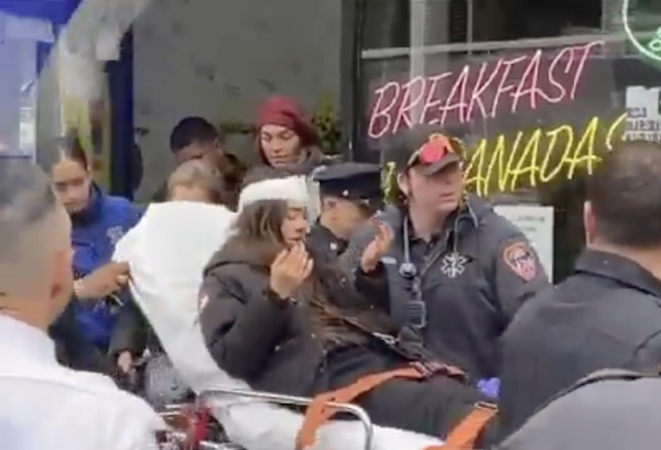 A young girl with long brown hair lies on a stretcher as first responders wheel her down a sidewalk filled with people. The girl is strapped down and has white bandages around her head.