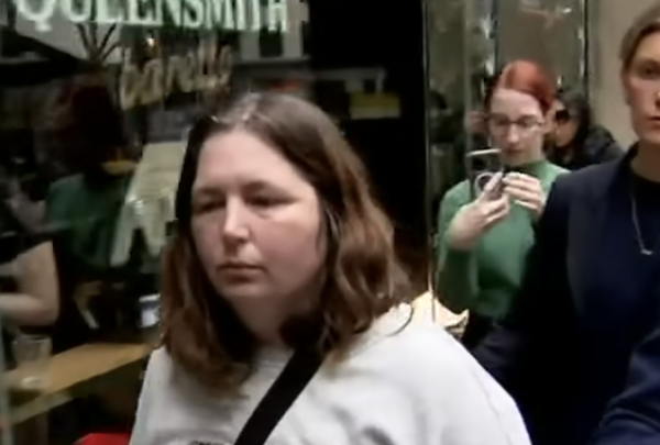 A white woman with shoulder-length straggling brown hair walks down an urban street while a crowd films her.