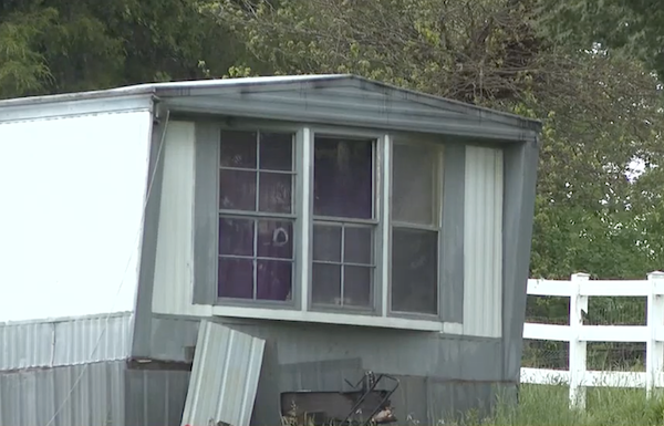 Daytime shot of one end of a white single-wide mobile home with gray trim. There are three windows visible on the end of the home and one has a large hole in it. The trailer sits in a green field beside a white fence and looks older and in slight disrepair.