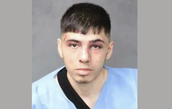 A young man with short dark brown hair has bruises and cuts on his face and stares expressionless during his mugshot. He wears a light blue shirt and has a black strap draped over one shoulder.