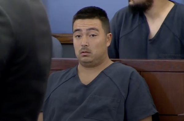 A man with short dark brown hair and facial stubble looks surprised as he sits in a navy blue jail issued shirt inside a courtroom with blue walls.