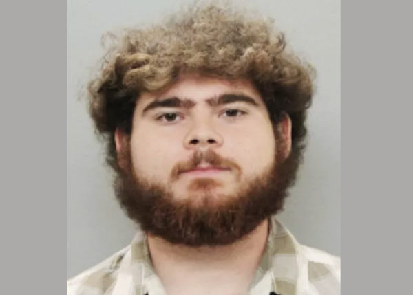 A young white man with short wildly curly and frizzed out brown hair has a full brown beard and wears a gray and white collared shirt as he stares expressionless during his mugshot.