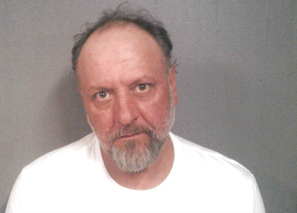 An older white man with receding brown and gray hair and a full gray beard tilts his head and frowns slightly at the camera during his mugshot. He wears a white t-shirt.